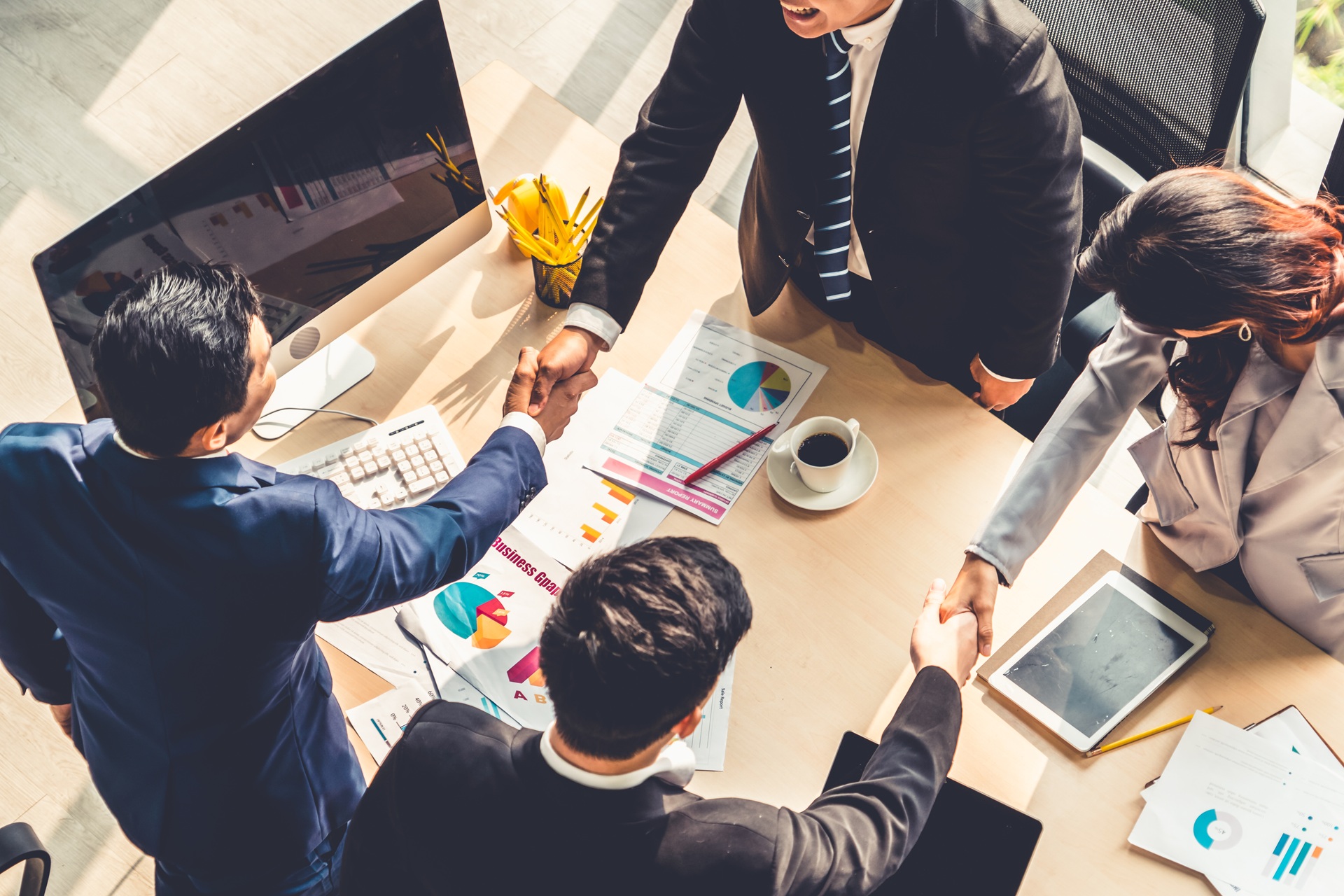A group of business people shaking hands over a table covered with electronic devices and reports.